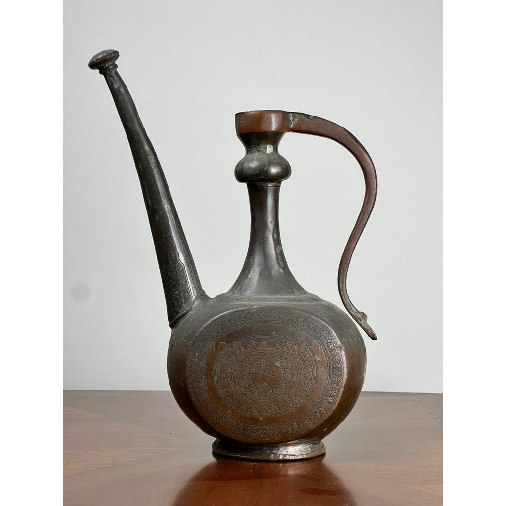 An Antique Qajar Persian Copper Engraved Ewer (19th Century) with a long spout, rounded base, and ornate patterns is displayed on a wooden surface against a plain light background. Brand: Unknown.