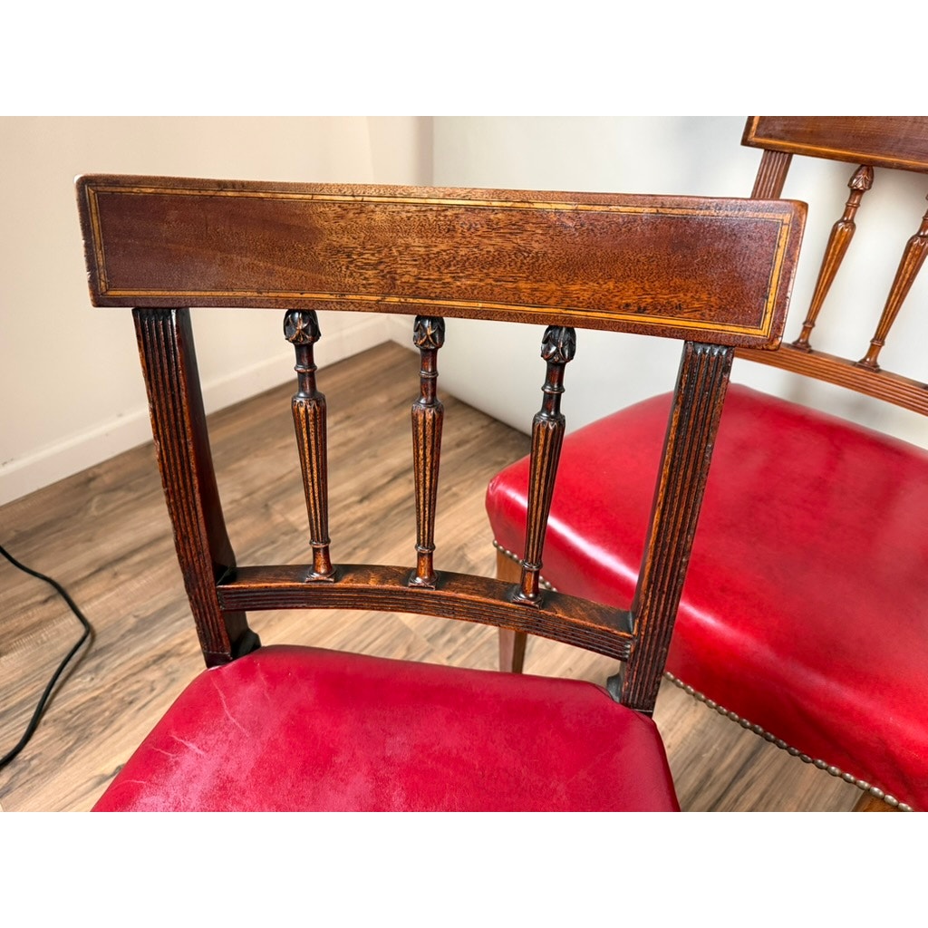 A set of six elegant English George III mahogany neoclassical dining chairs—four from the 18th century—by Pearl City Antiques, featuring decorative carved backrests and red upholstered seats, are arranged on a wooden floor near a cream-colored wall.