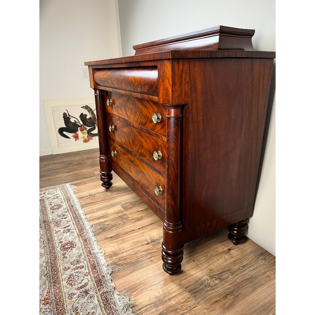 An Antique American Empire Albany Four Drawer Chest c. 1840 stands on a wood floor by a decorative rug; ornate brass handles and carved legs add charm, while framed artwork rests against the wall in the background.