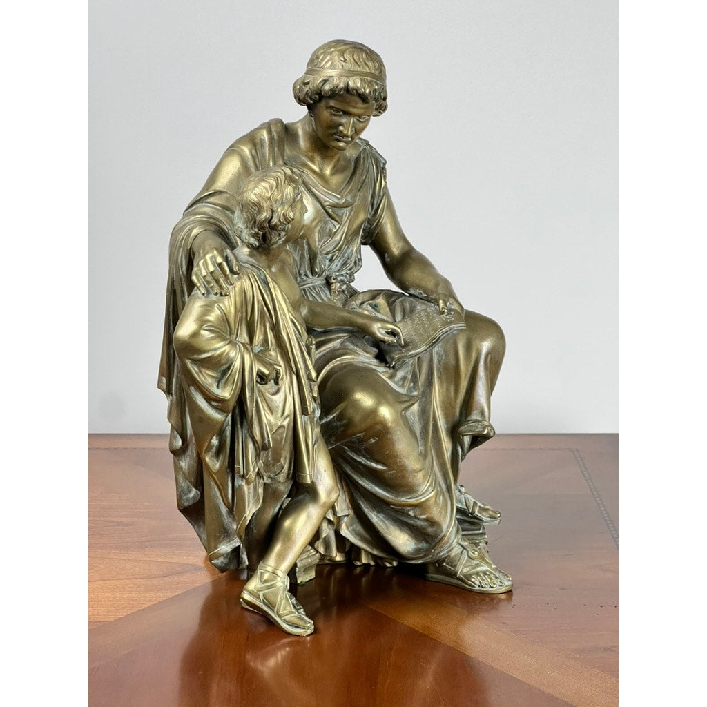 The Antique Bronze Sculpture “Teacher and Pupil” by Louis Charles Janson (1865, signed Ch. Janson) depicts a seated woman reading to a child in classical robes, displayed on a polished wood surface against a plain background.