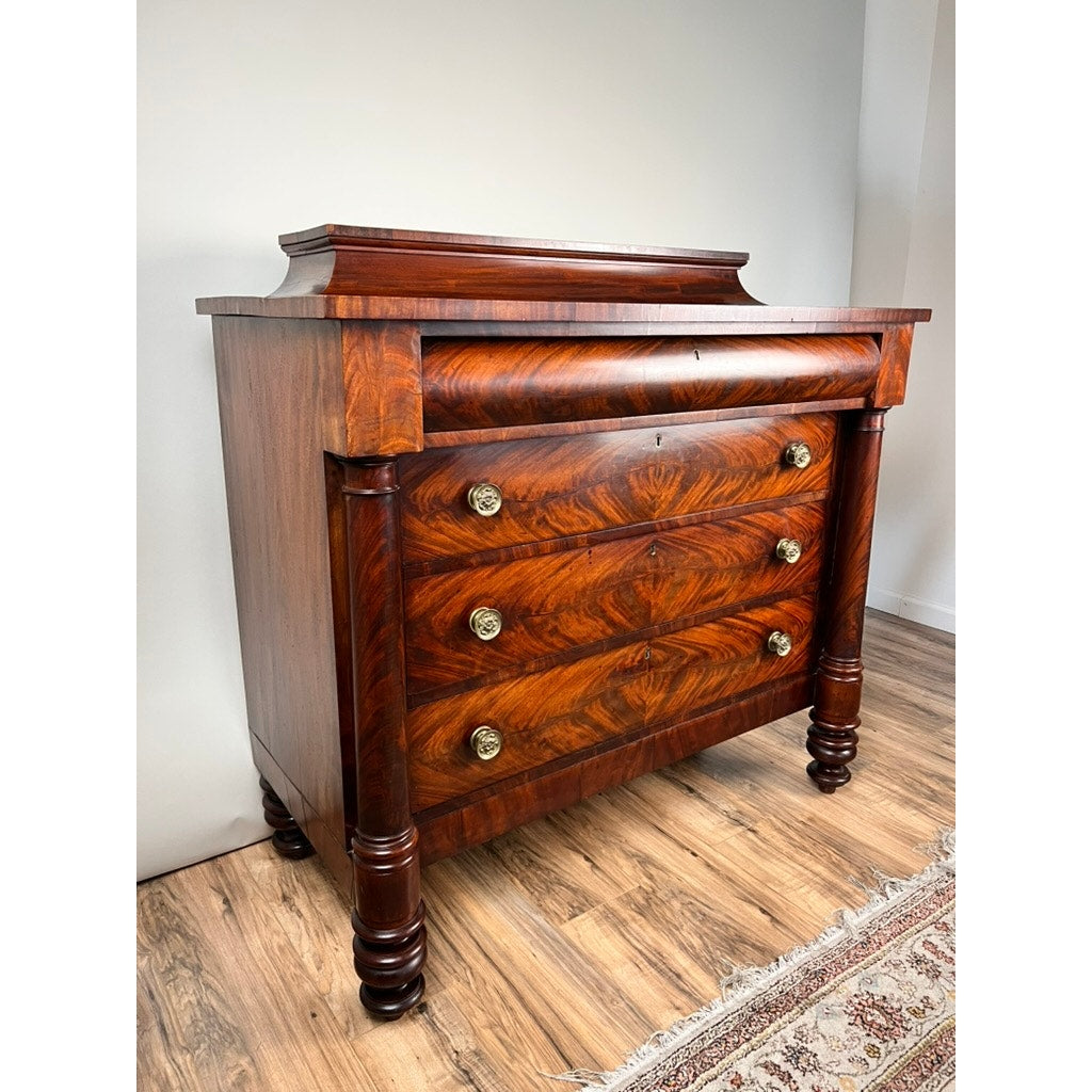 The Antique American Empire Albany Four Drawer Chest c. 1840, featuring ornate brass handles and rich flame mahogany grain with carved legs, stands on a wooden floor beside a patterned rug and plain white wall.