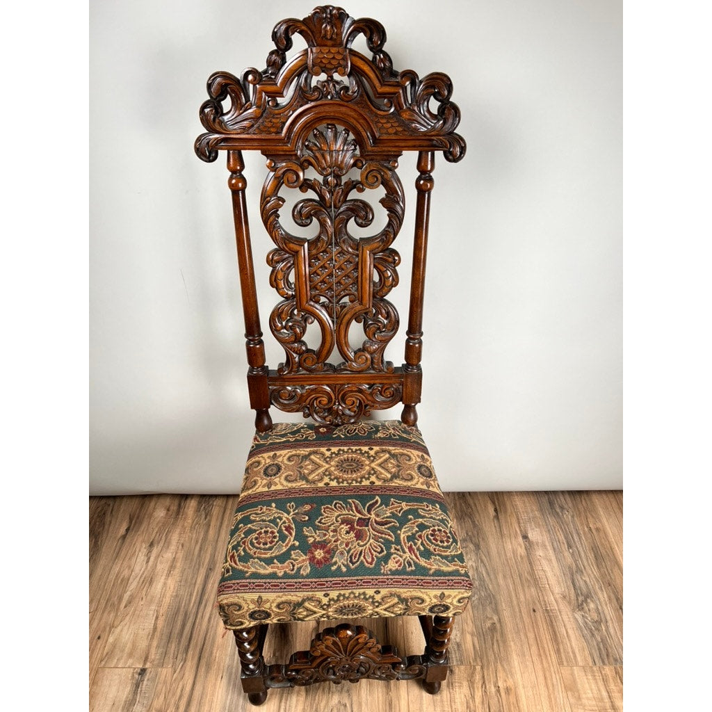 The Antique Carolean Revival Side Chair, c. Late 19th Century, has an ornate dark wood frame, intricate carvings on the tall back and legs, and a patterned green, gold, and red upholstered seat—displayed on a wooden floor against a plain wall.
