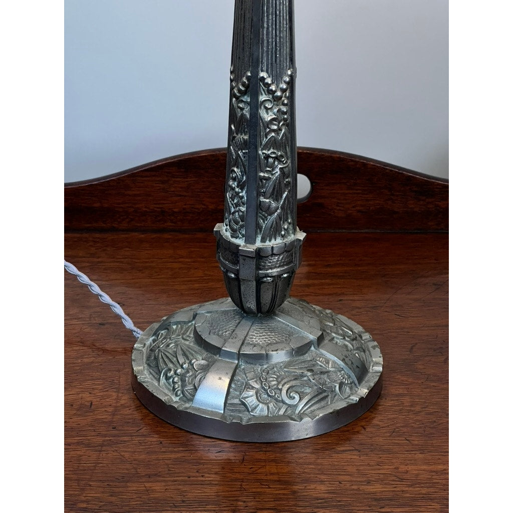 The image features the ornate metal base of a Vintage French Art Deco Frosted Glass Table Lamp by Muller Frères Lunéville c. 1920, with detailed floral and leaf patterns and a twisted white cord, set on a polished wooden surface.