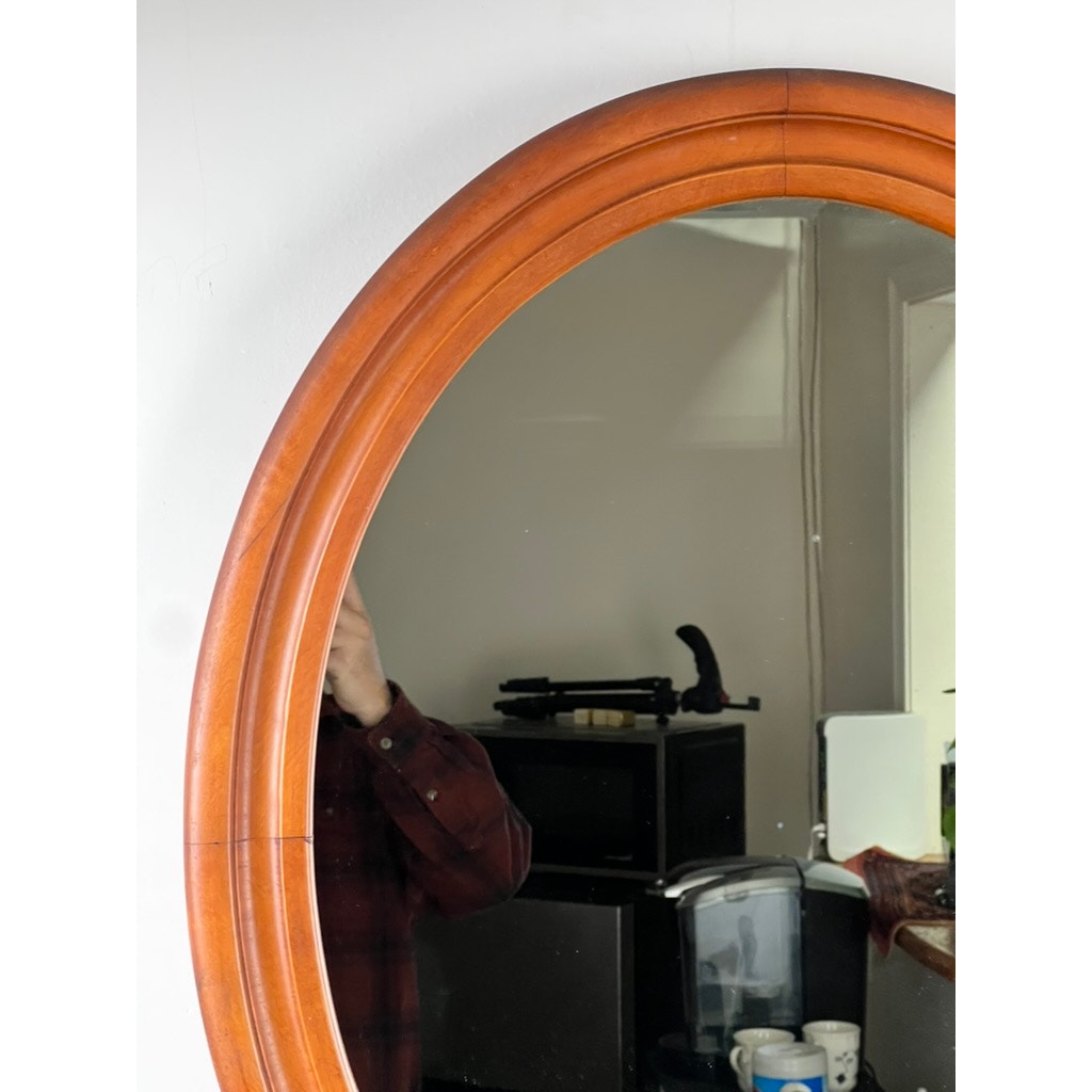 A person in a red plaid shirt is partially visible in the reflection of a Vintage Oval Maple Wall Mirror, with kitchen appliances, mugs, a black coffee maker, and a white wall behind them. Brand: None/Unknown.