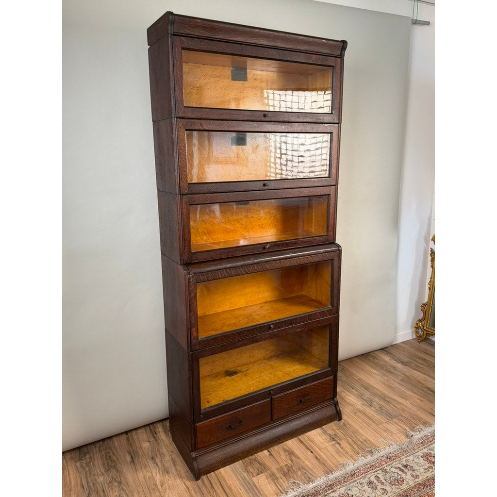 The Antique Hale System Quarter-Sawn Oak Five-Stack Barrister Bookcase by Camden Cabinet Company (c. 1910) stands on a hardwood floor near a white wall, featuring five glass-fronted shelves and two small drawers at the bottom, with a partial view of a rug nearby.