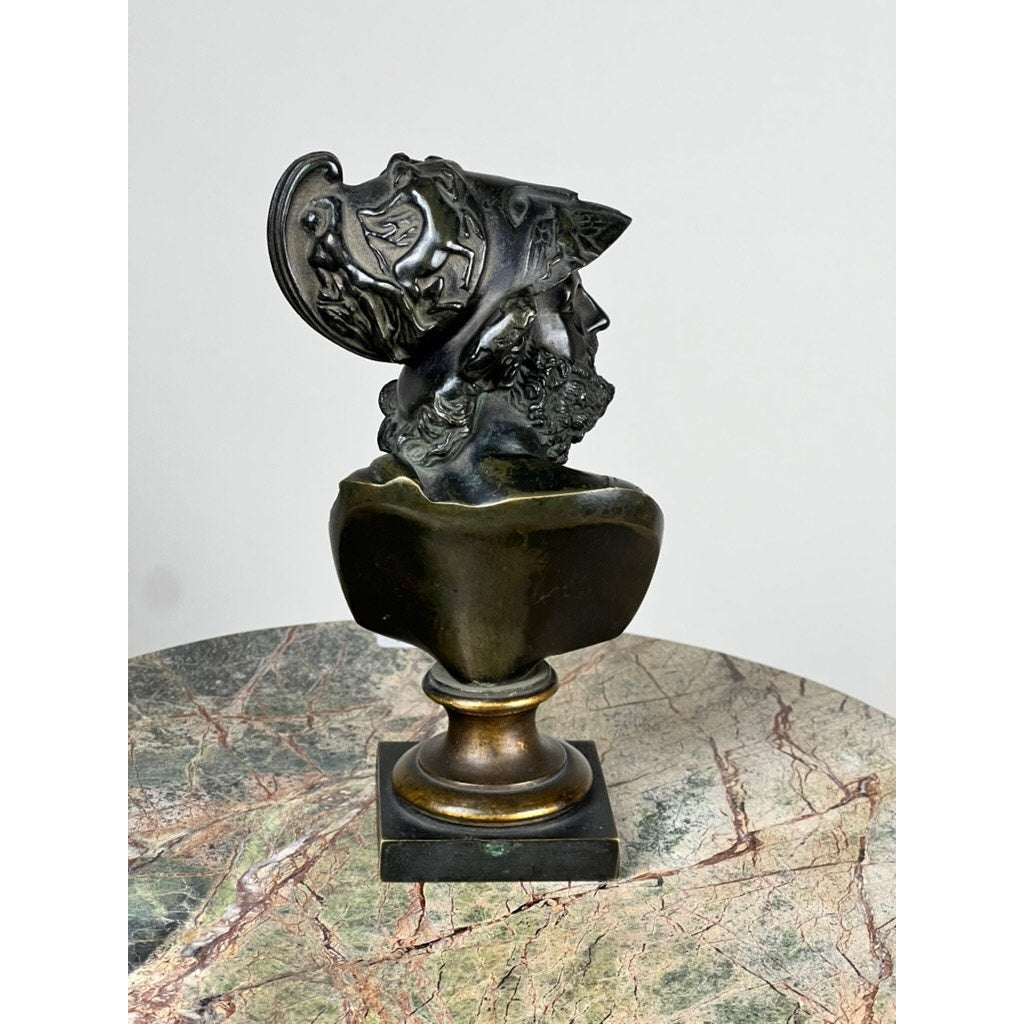 Antique Small Bronze Bust of King Menelaus, late 19th century, featuring an ornate helmet with intricate carvings. The bust is mounted on a pedestal and displayed on a marble-like surface against a plain light background. Brand: Unknown.