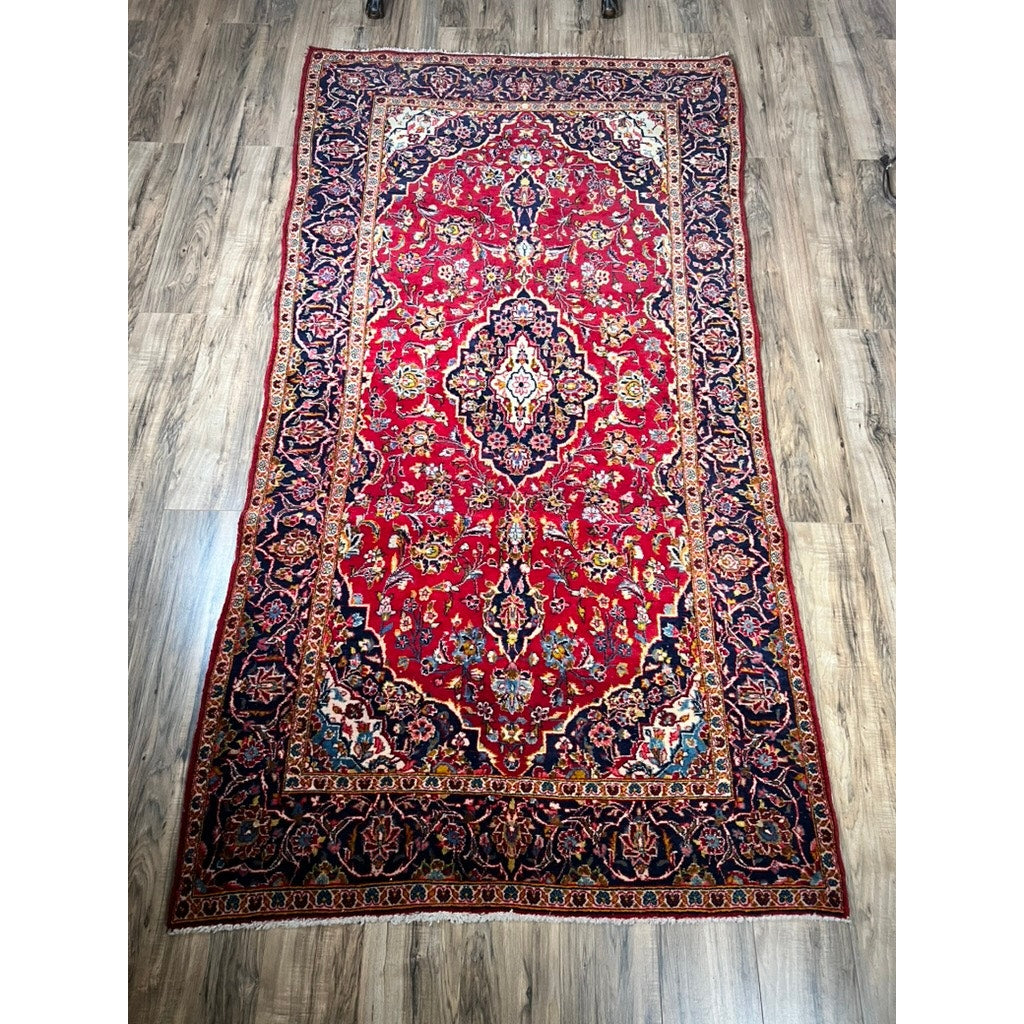 A vintage hand-knotted Persian Kashan wool gallery rug (c. 1950–1970, 4’5 x 8’8), featuring a red center, intricate floral designs, and a dark blue ornate border, is displayed on a wood floor.