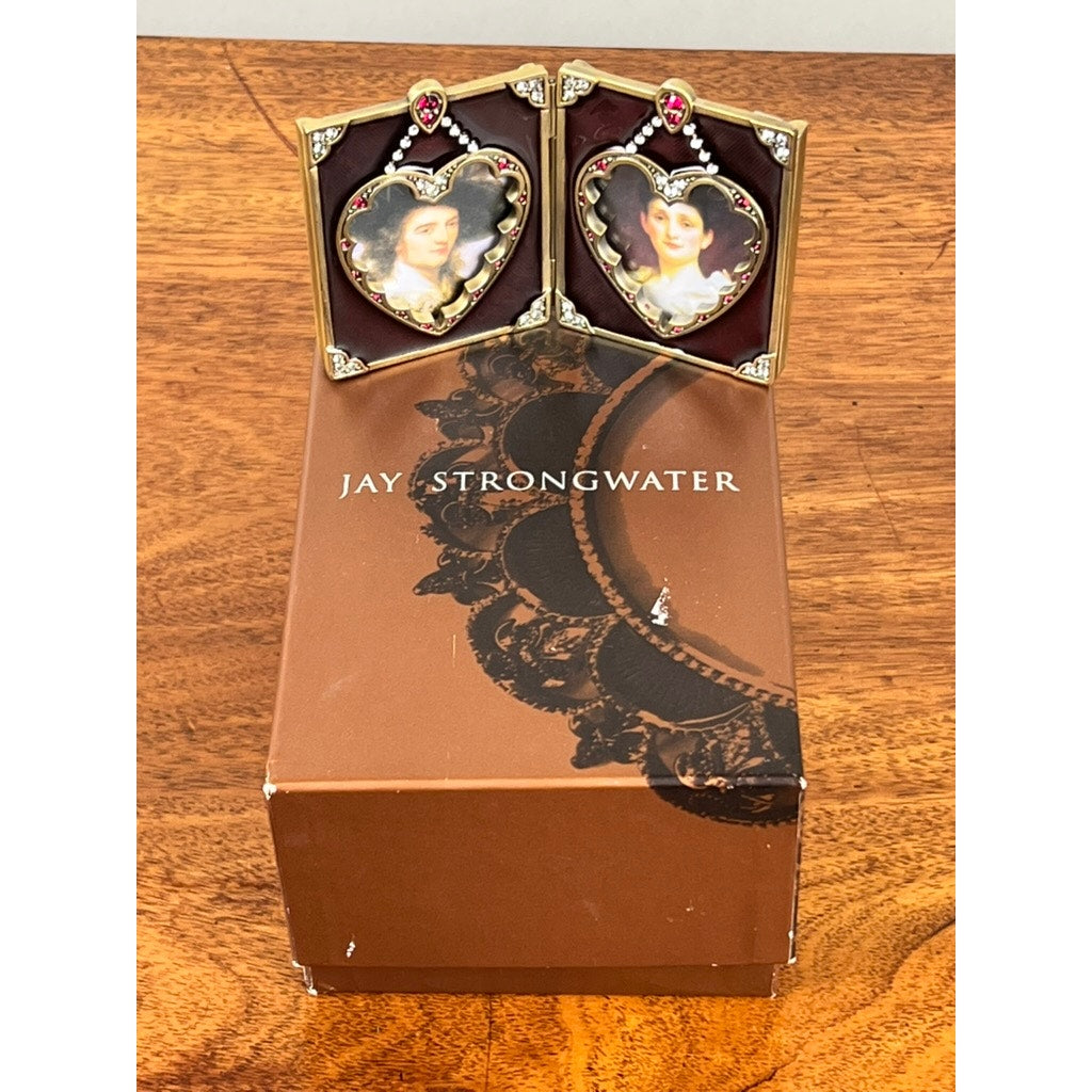 A new Jay Strongwater Bejeweled Enamel Double Heart Picture Frame, featuring jeweled accents and displaying portraits of a man and woman, is shown on a brown wooden surface.
