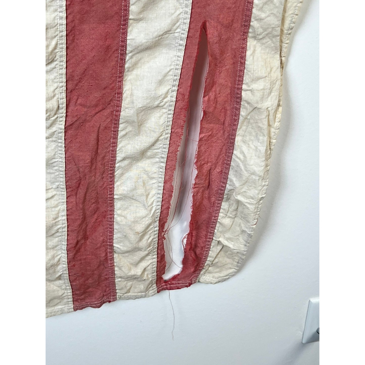 A close-up of a vintage Valley Forge “76” Bennington American Flag from the Bicentennial Era (c. 1976), featuring worn red and white stripes with a significant vertical tear along one of the white stripes, revealing the wall behind it. Brand is unknown.