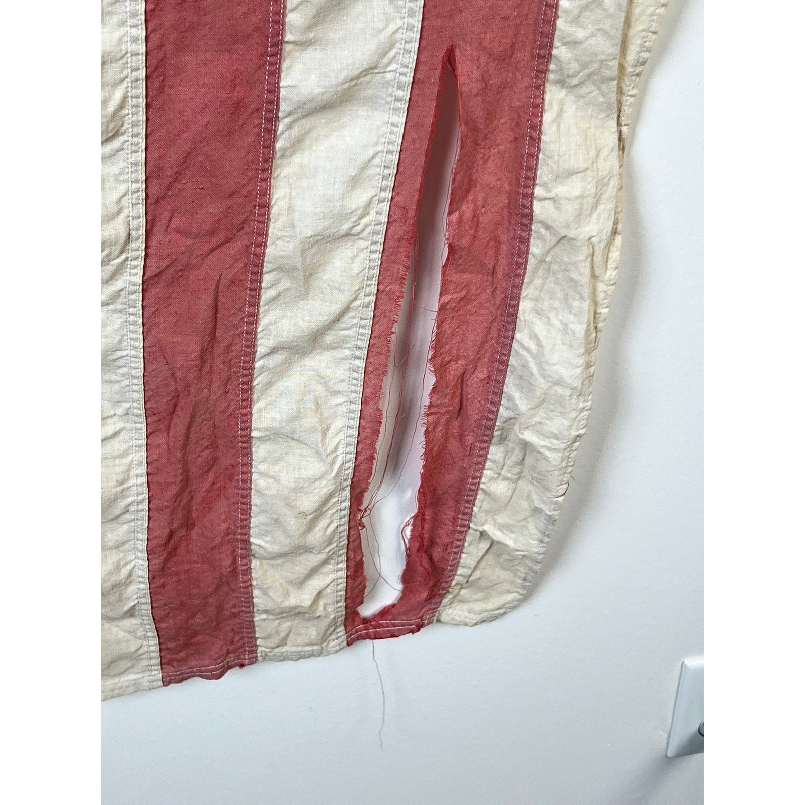A close-up of a vintage Valley Forge “76” Bennington American Flag from the Bicentennial Era (c. 1976), featuring worn red and white stripes with a significant vertical tear along one of the white stripes, revealing the wall behind it. Brand is unknown.