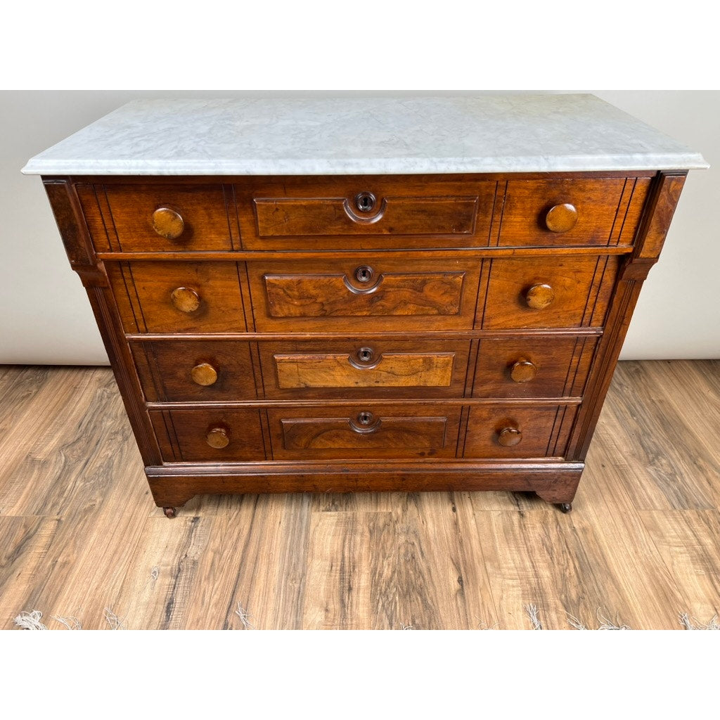 An Antique Victorian Marble Top Four Drawer Chest c. 1910 with a white marble top and classic wooden knobs stands elegantly on a wooden floor.