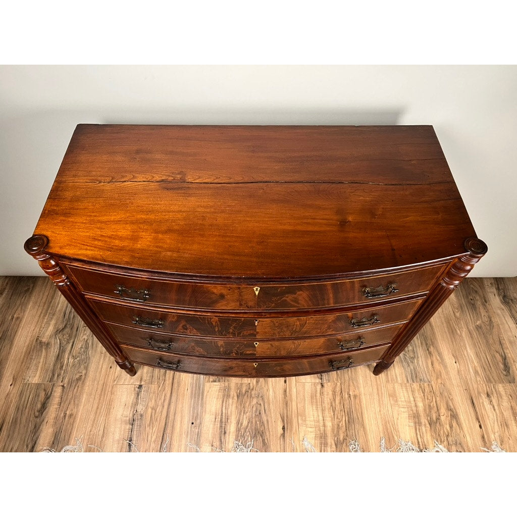 A top-down view of an Antique Sheraton Mahogany Bowfront Chest (early 19th century) with four drawers, ornate metal handles, and a polished finish, placed on a wooden floor by a fringed rug. Brand: Unknown.