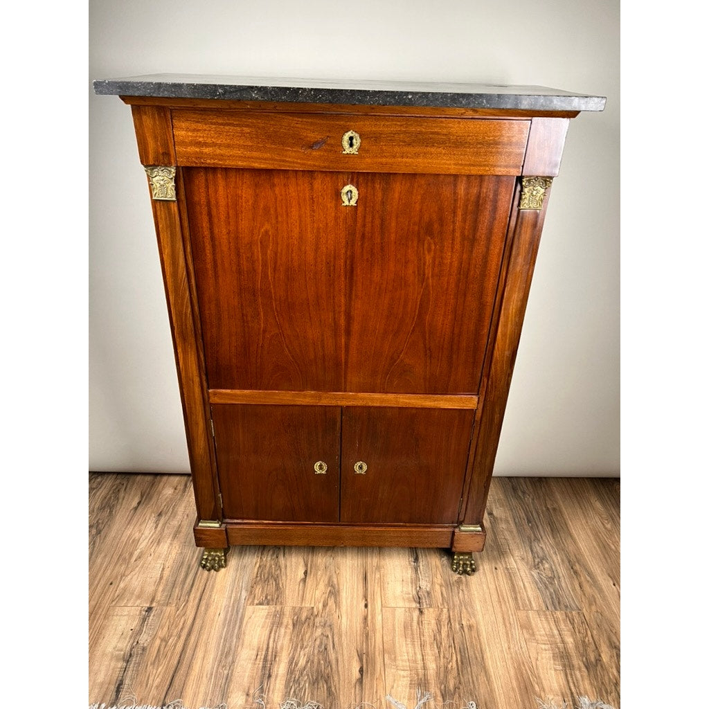 An Antique French Empire Secrétaire Abattant with ormolu mounts (mid 19th century) stands on a wooden floor against a light-colored wall, featuring a dark marble top and gold accents on corners, feet, and keyholes. Brand: Unknown.