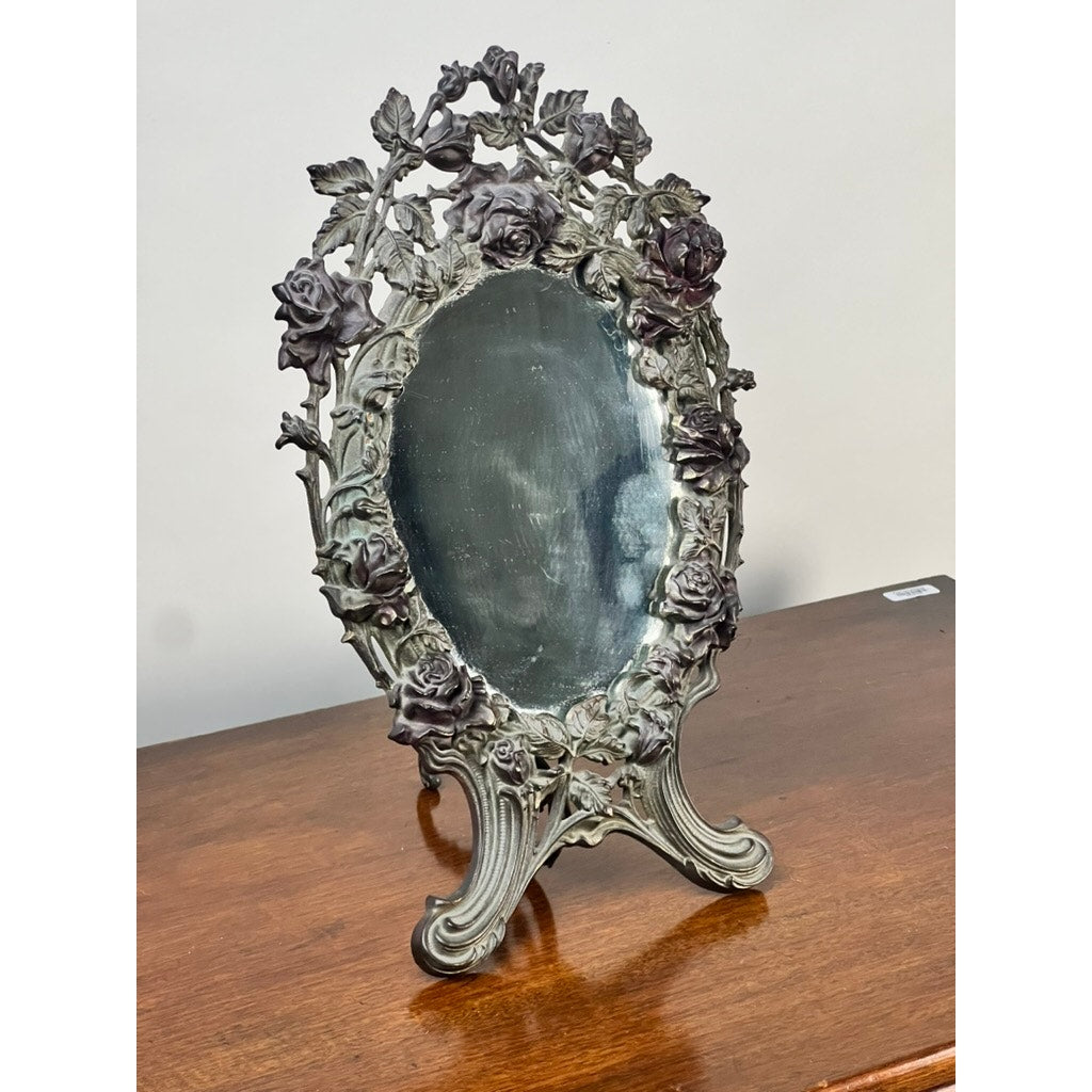 An Antique Art Nouveau Silvered Cast Brass Rose Vanity Dressing Mirror, dated 1909, with an ornate floral frame and stand, rests on a polished wooden surface against a plain light background. Brand is unknown.