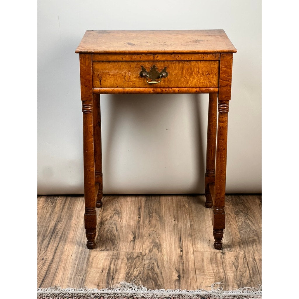 An Antique Sheraton Birdseye Maple One Drawer Stand from the early 19th century, featuring slender turned legs and a brass-handled drawer, stands on a wood-patterned floor against a plain light wall.