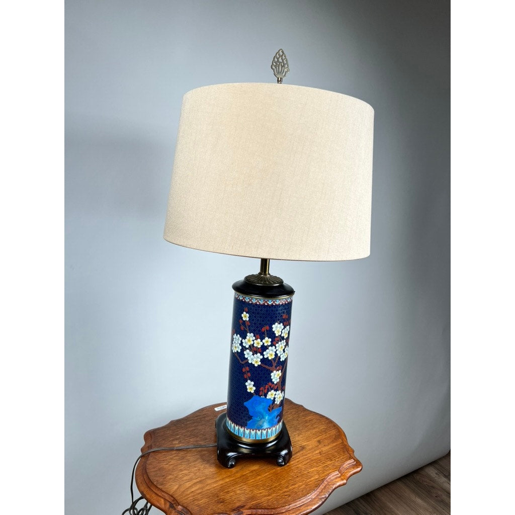 The Vintage Japanese Cloisonné Cylinder Table Lamp by Knob Creek features a blue floral ceramic base and cream shade, shown on a wooden side table against a plain light gray background.