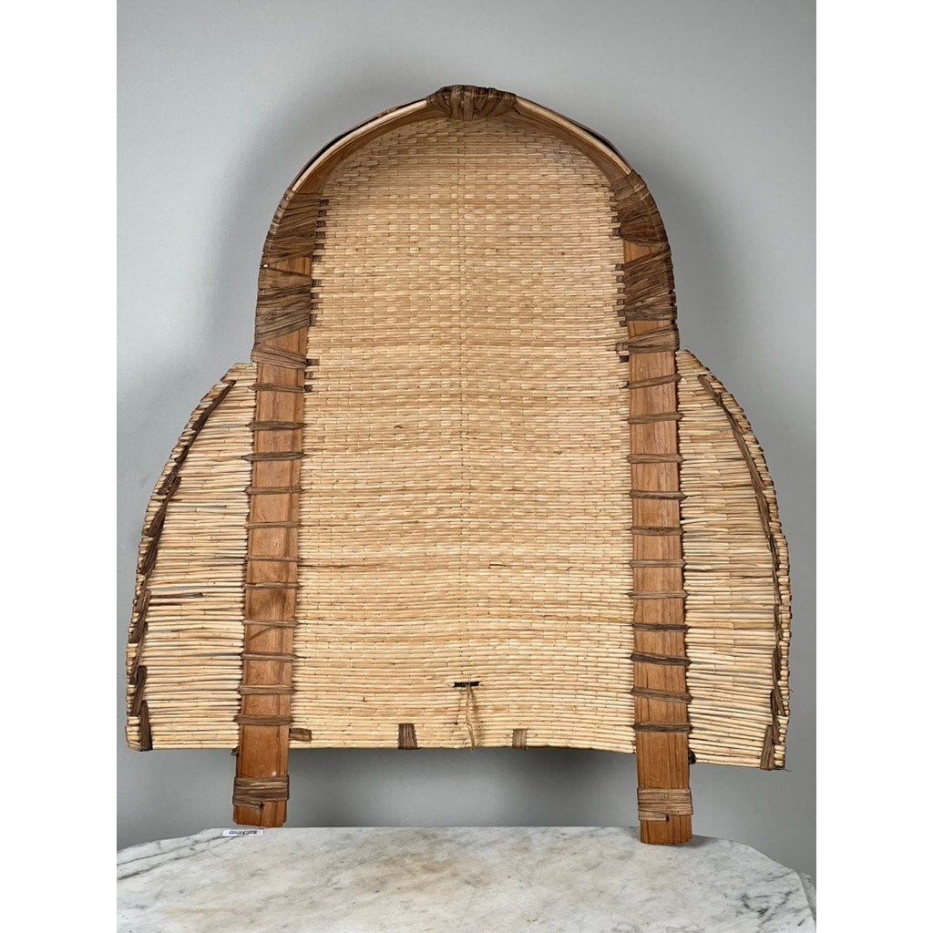 The Vintage Traditional Winnowing Basket, a woven dome-shaped piece made from natural fibers with two short legs, is displayed against a plain light gray wall and set atop a white marble surface.