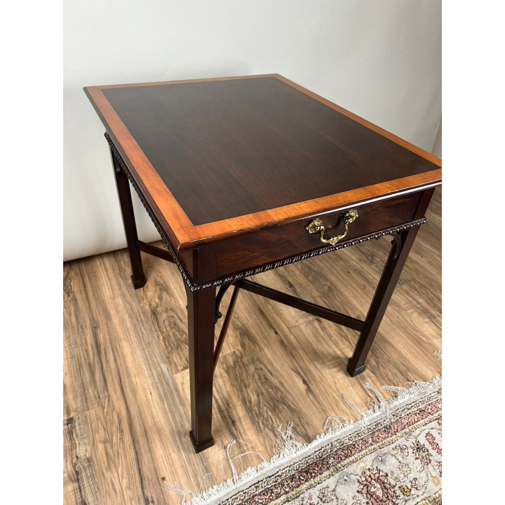 The Stickley Vintage Federal Style X Stretcher One Drawer End Table features a contrasting wood border, brass handle, and decorative trim, standing on a wooden floor beside a patterned rug.