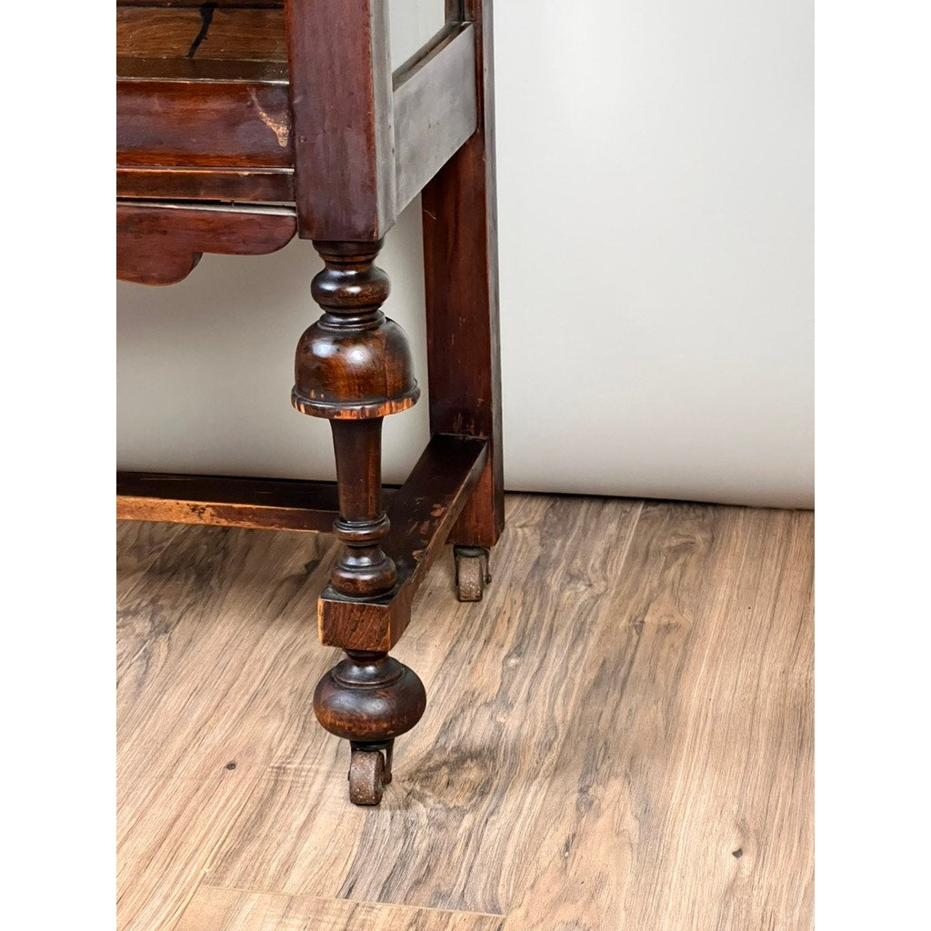 A close-up of a carved mahogany leg with a small caster wheel, part of a Depression Era Mahogany Curio Display Cabinet with Glass Sides from the 1930s, standing on a light wood floor against a plain white wall.