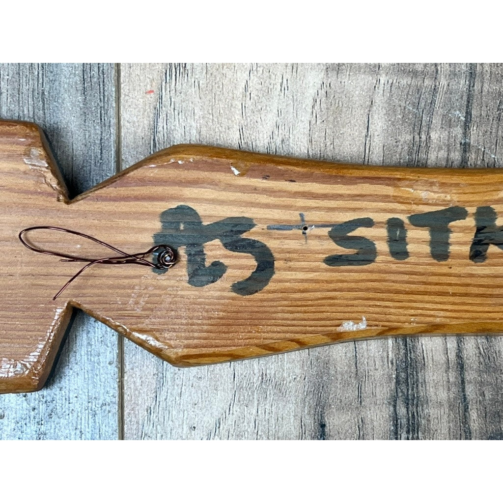 A close-up of a Vintage Inuit Carved Red Cedar Paddle from Sitka, Alaska, signed illegibly, shows partially visible painted text with “SIT” and a plus sign. A wire loops through a hole on the left as it rests on a gray wooden surface.