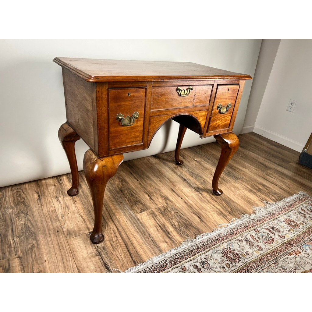 An Antique George III Queen Anne Mahogany Lowboy, late 18th century, with three drawers and brass handles, features curved cabriole legs and stands on a hardwood floor beside a patterned rug and white wall.