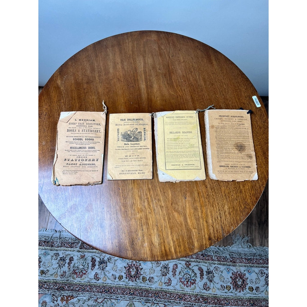 An antique set of four old Farmer’s Almanacs (1858, 1861, 1864 & 1865 by Robert B. Thomas) with yellowed covers are laid side by side on a round wooden table, with a patterned rug partially visible beneath.