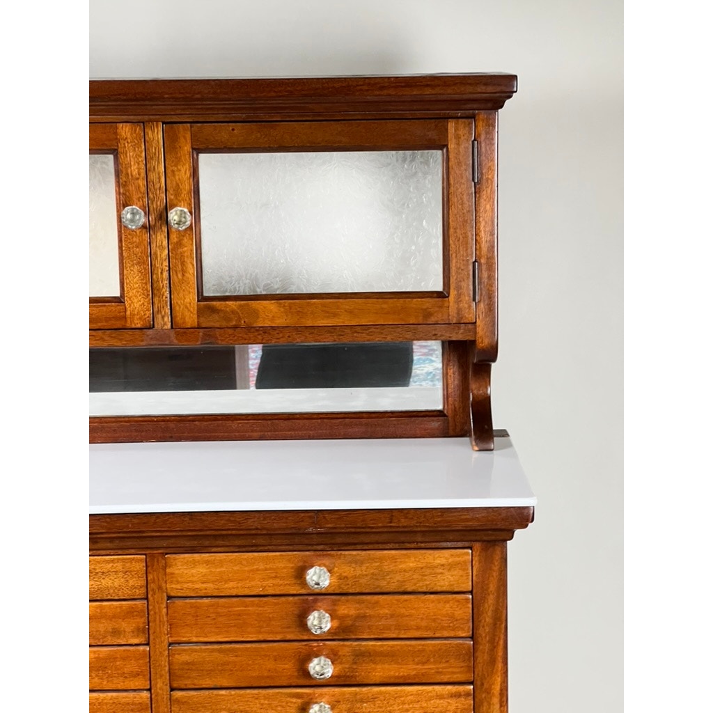 A partial view of an Antique Mahogany Multi-drawer Dental Cabinet by The American Cabinet Co., early 20th century, shows frosted glass upper doors, a clear lower panel, and multiple drawers with round metal knobs against a plain light wall.