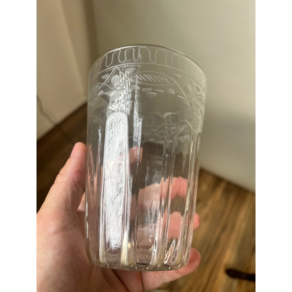 A hand holds the Attributed Boston & Sandwich Wheel Engraved Mold-Blown Glass Table Vase, Circa 1845–1860—its clear form adorned with intricate engraved patterns near the rim—against a softly blurred indoor background with wooden flooring.