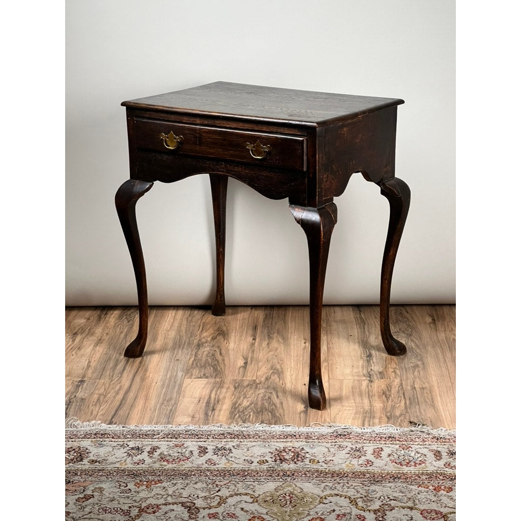 An Antique Queen Anne Style George III Oak One Drawer Stand (late 18th century) stands on a wooden floor beside a patterned rug and against a plain light-colored wall.