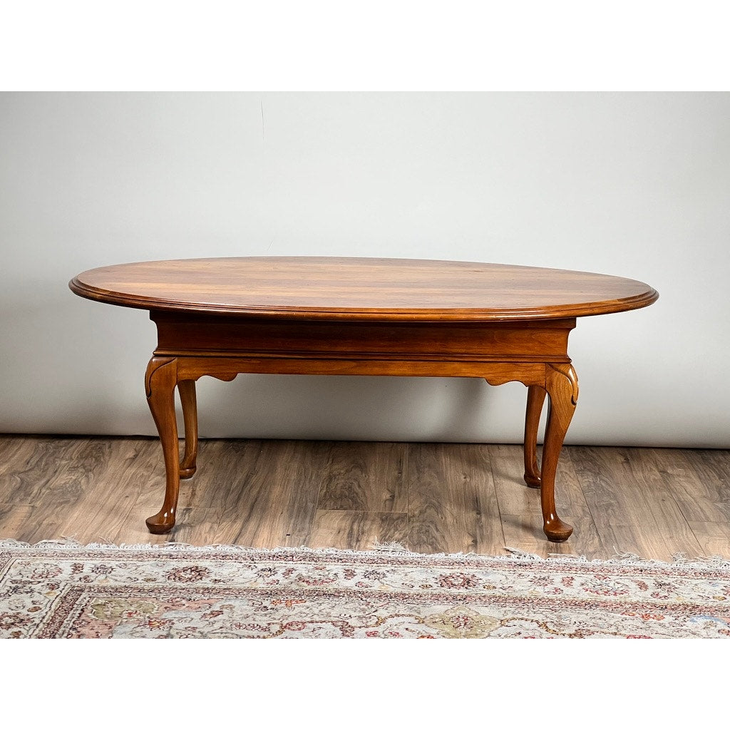 A Vintage Solid Cherry Queen Anne Style Coffee Table, attributed to Pennsylvania House, with an oval shape and curved legs rests on a hardwood floor near a patterned rug against a light-colored wall.