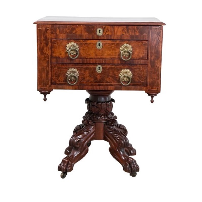 An American Classical Figured Mahogany Work Table, circa 1830–1845, featuring two drawers with brass lion-head ring pulls and an ornate pedestal base with carved feet on casters.