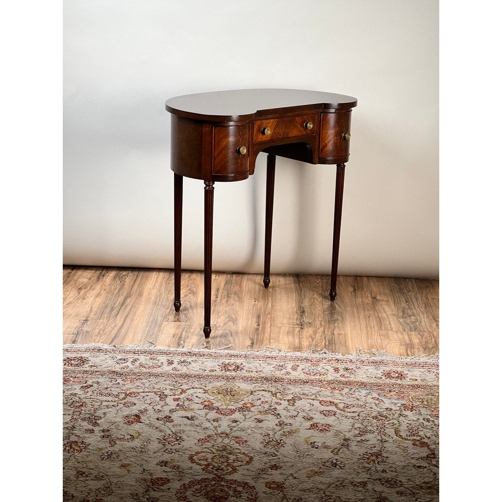 A Vintage Edwardian Sheraton Revival kidney-form dressing table c. 1940 with mahogany veneers, three brass-handled drawers, curved edges, and slender legs stands on a hardwood floor by a patterned rug and light-colored wall. Brand: Unknown.