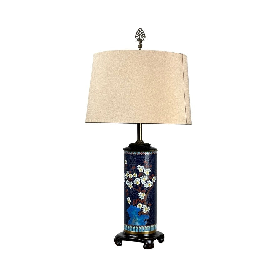 The Vintage Japanese Cloisonné Cylinder Table Lamp by Knob Creek features a beige shade, dark blue ceramic base with floral and bird motifs, and rests on a decorative black stand—evoking classic vintage style.