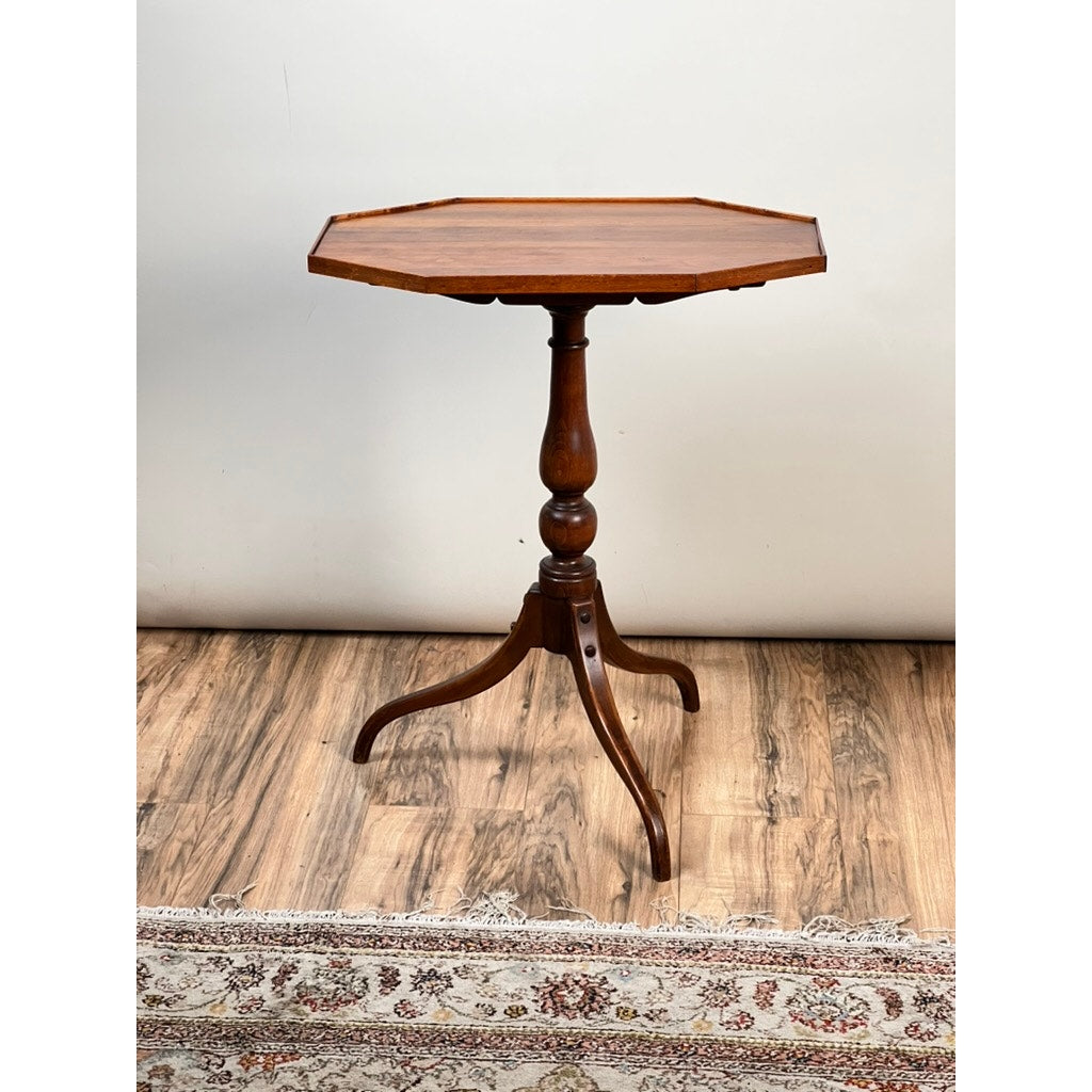 A Vintage Federal Style Cherry Octagonal Tilt Top Candlestand by Stickley with a turned pedestal and three curved legs stands on a wooden floor partly covered by a patterned rug.