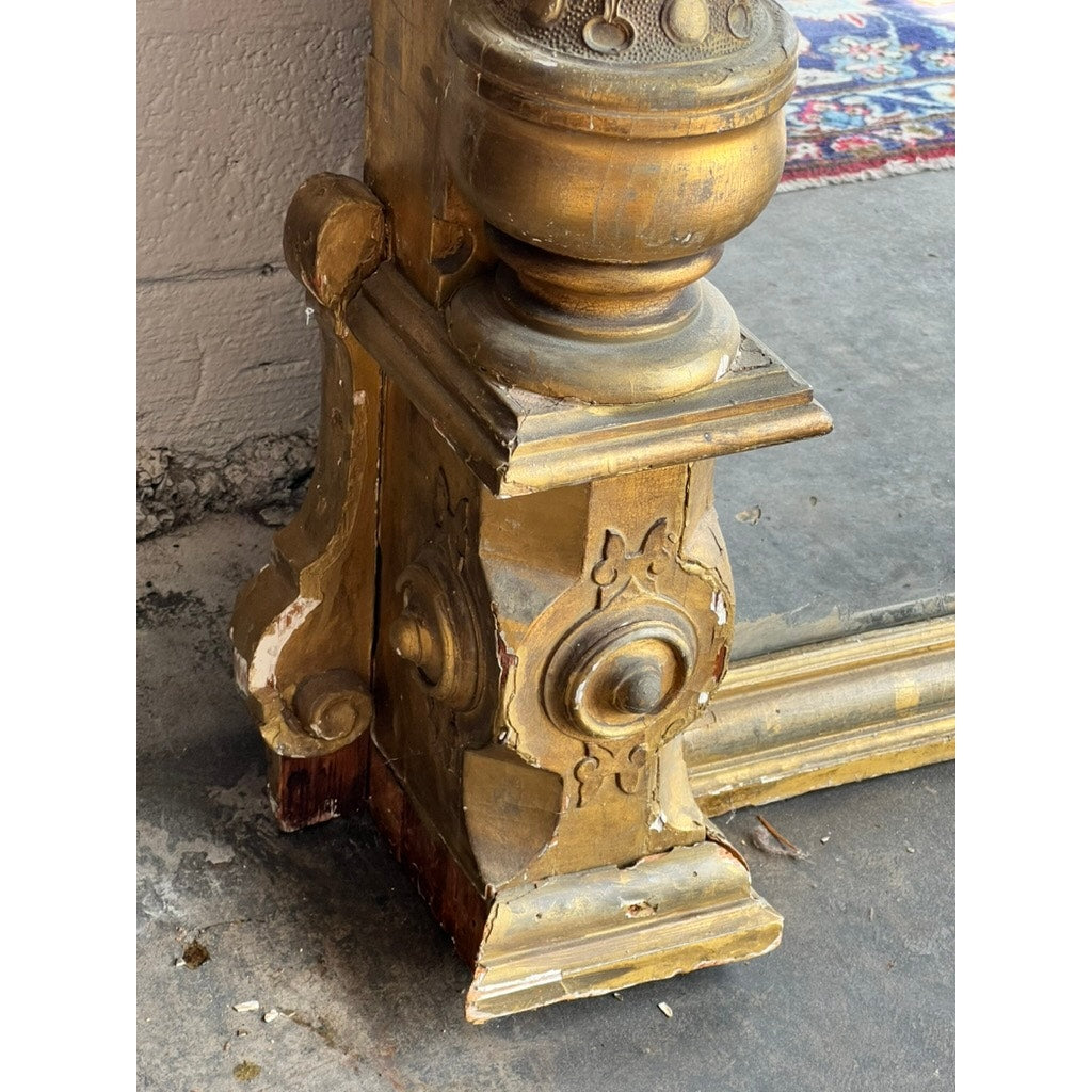 Close-up of an ornate, gold-painted giltwood frame with worn and chipped areas, adjacent to an Exceptional Monumental Victorian Renaissance Revival Pier Mirror, circa 1870–1885. A section of a colorful rug can be seen in the background on the concrete floor, highlighting the mirror’s timeless elegance.