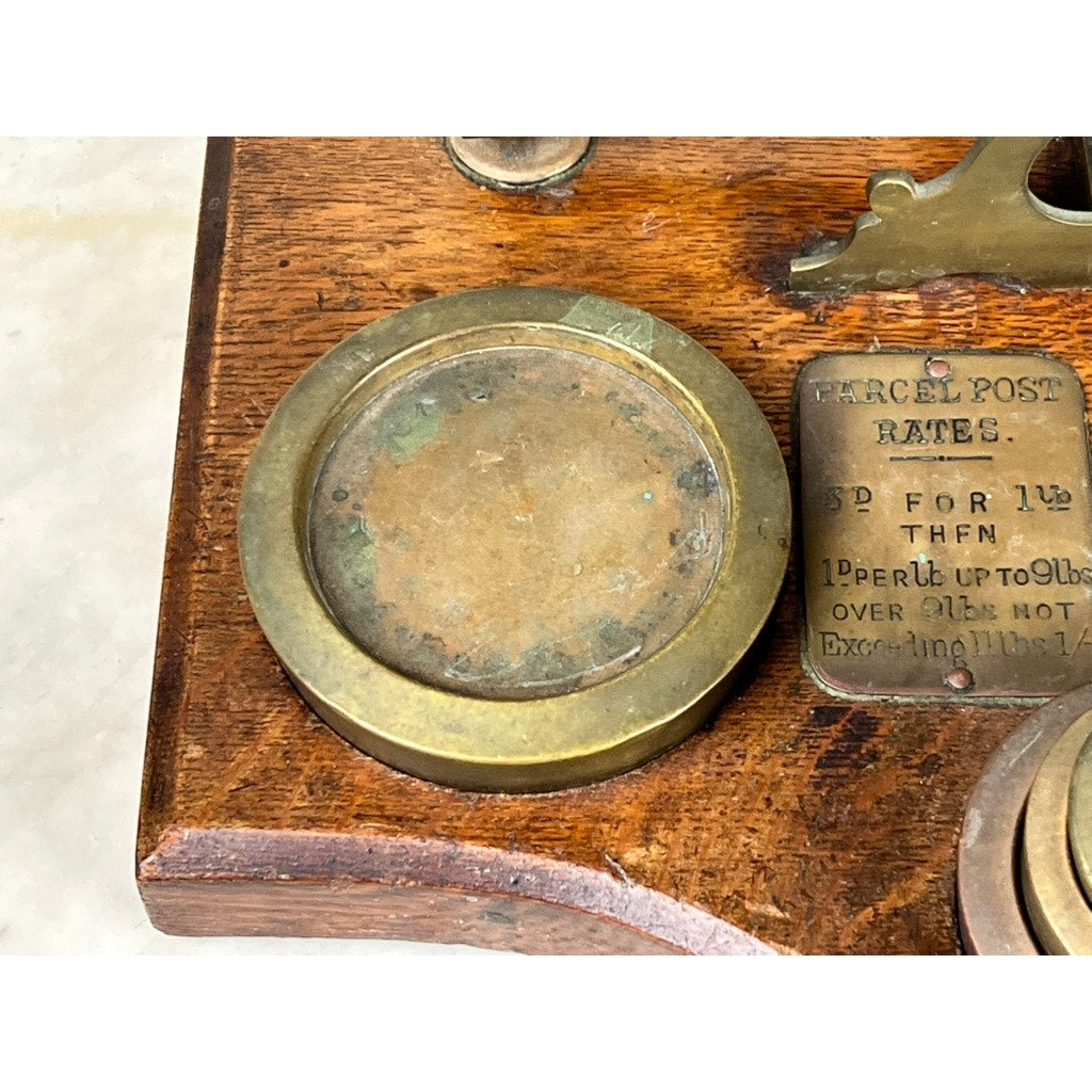 A close-up of the Antique 19th Century English Postal Scale on a wooden base, beside a small metal plaque listing parcel post rates and postal details. Brand: None/Unknown.