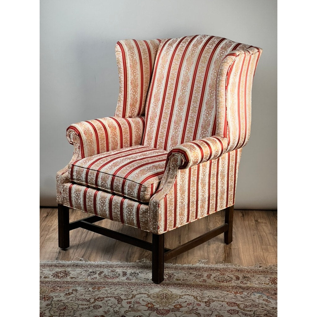 A Vintage Chippendale Style Wingback Chair by Ethan Allen features traditional red and cream striped floral upholstery, wooden legs, and sits on a wooden floor next to a patterned rug.