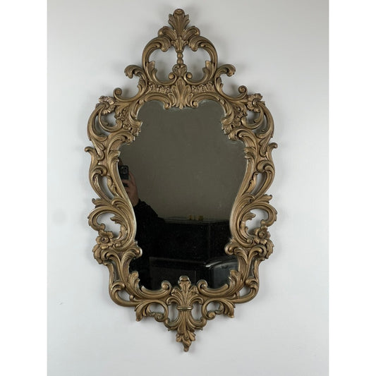 Vintage Rococo Style Decorators Mirror by Syracuse Ornamental from Pearl City Antiques, featuring an elaborate scrollwork frame, reflects part of a person holding a phone and some furniture against a plain white wall.