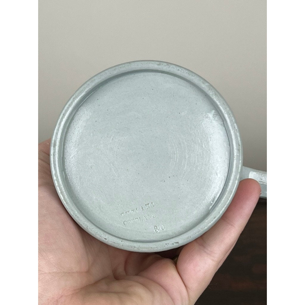 A hand holds a Vintage Copeland Spode English Jasperware Fox Hunt Pitcher upside down, displaying its smooth base with faint makers marks and numbers. The softly blurred background features a beige wall and dark surface. Brand: Copeland Spode.