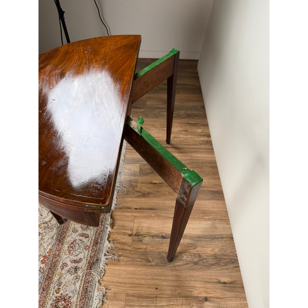 An English George III Mahogany and Satinwood-Inlaid Demilune Games Table featuring a mahogany hinged top, an extendable rear leg mechanism, and a green fabric lining.