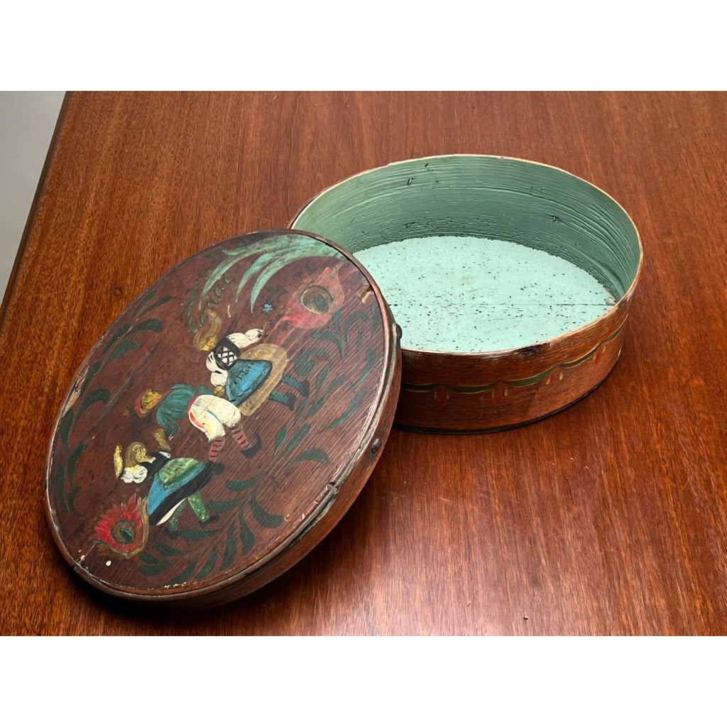 An Antique German polychrome painted bentwood "Spanschachtel" ribbon box (late 18th century) with a lid showing two figures in traditional dress among flowers; open to display its pale green interior on a polished wood surface. Brand unknown.