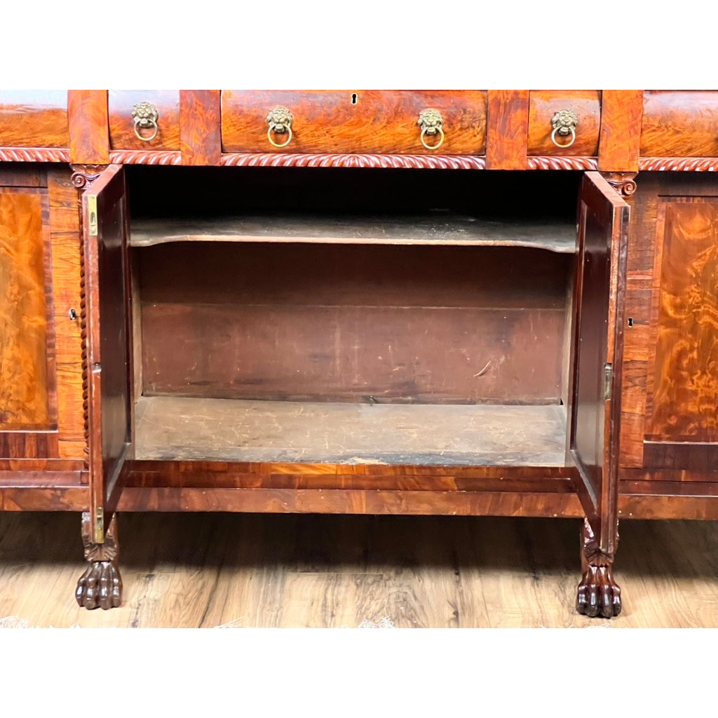 An Antique American Empire Mahogany Sideboard with rosewood edge banding, ornate flame mahogany grain, open doors, empty shelf, clawed feet, decorative brass pulls, and carved details stands on a wood floor. Likely Philadelphia c. 1840.