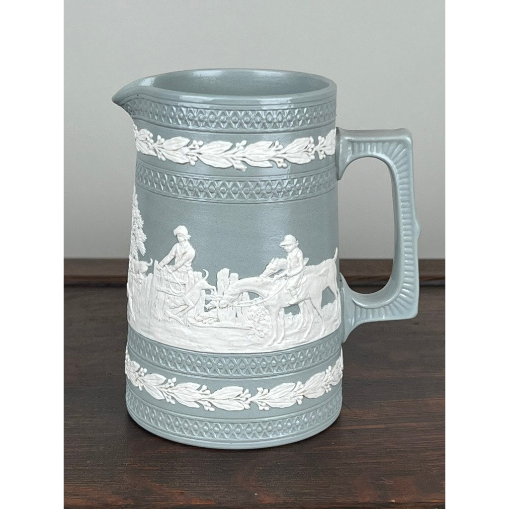 A Vintage Copeland Spode English Jasperware Fox Hunt Pitcher features light blue jasperware with white classical reliefs of people and animals in a fox hunt, plus decorative leaf bands, displayed on a dark wood surface against a neutral background.