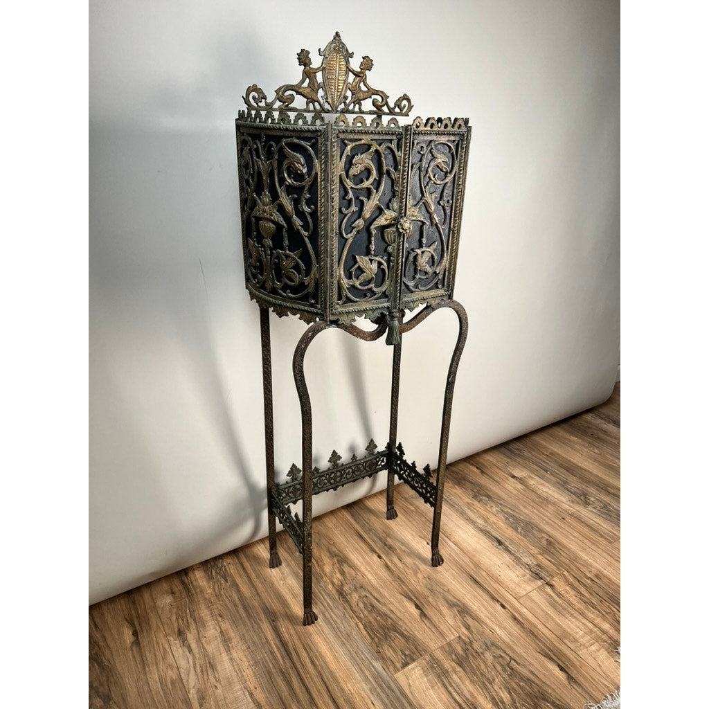 An Antique Gothic Revival cast iron and brass phone stand, attributed to Oscar Bach or Bertram Segar c. 1920 Greenwich Village, stands on four slender legs against a light wall and wood floor. Brand: None/Unknown.