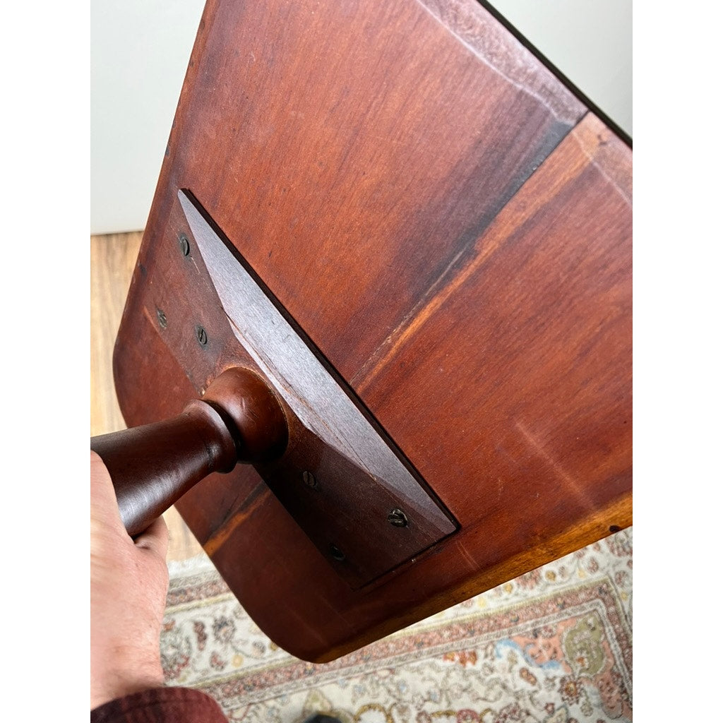 A close-up of a person holding the base of an Antique Federal Period Cherry Hepplewhite Candle Stand c Mid 19th Century, showing where the tabletop meets the pedestal, with the table above a patterned rug on a wooden floor.