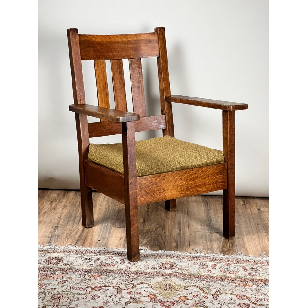 The Antique Quarter Sawn Mission Oak Arm Chair by Quaint Furniture c. 1920 features vertical slats, flat armrests, and a green upholstered seat cushion—an Arts & Crafts classic shown on a wooden floor with part of a patterned rug visible.