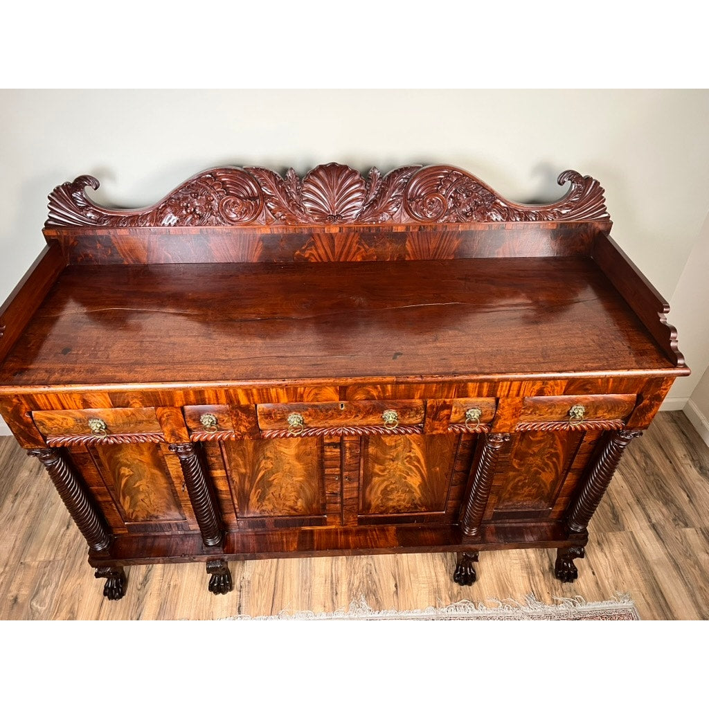 Antique American Empire Mahogany sideboard with rosewood edge banding, likely Philadelphia c.1840, featuring carved details, brass handles, fluted columns, claw feet, and an intricately carved back panel against a light wall and wood floor.