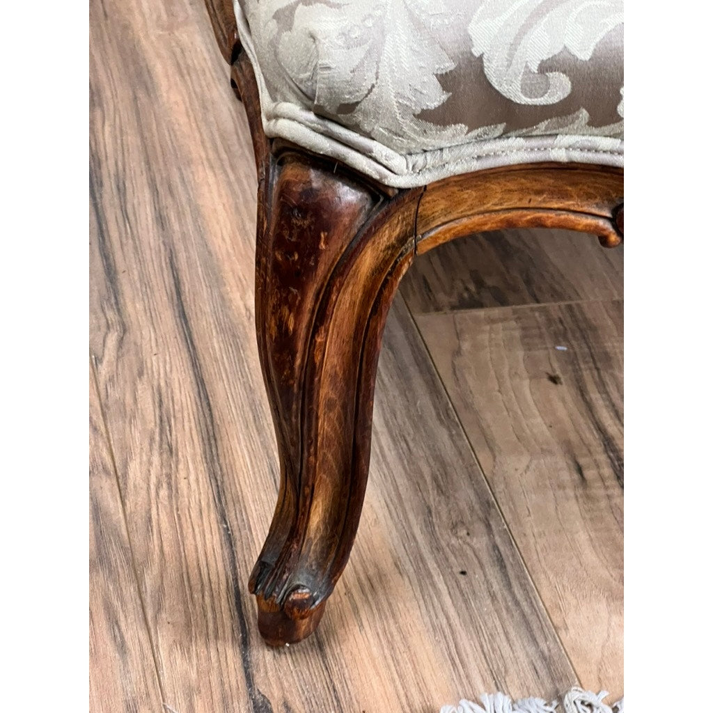 Close-up of an ornate carved walnut leg on an Antique French Louis XV Rococo Style Bergère Arm Chair, c. mid-19th century, featuring newer damask upholstery and a glimpse of fringe on the right, set on a wood floor.