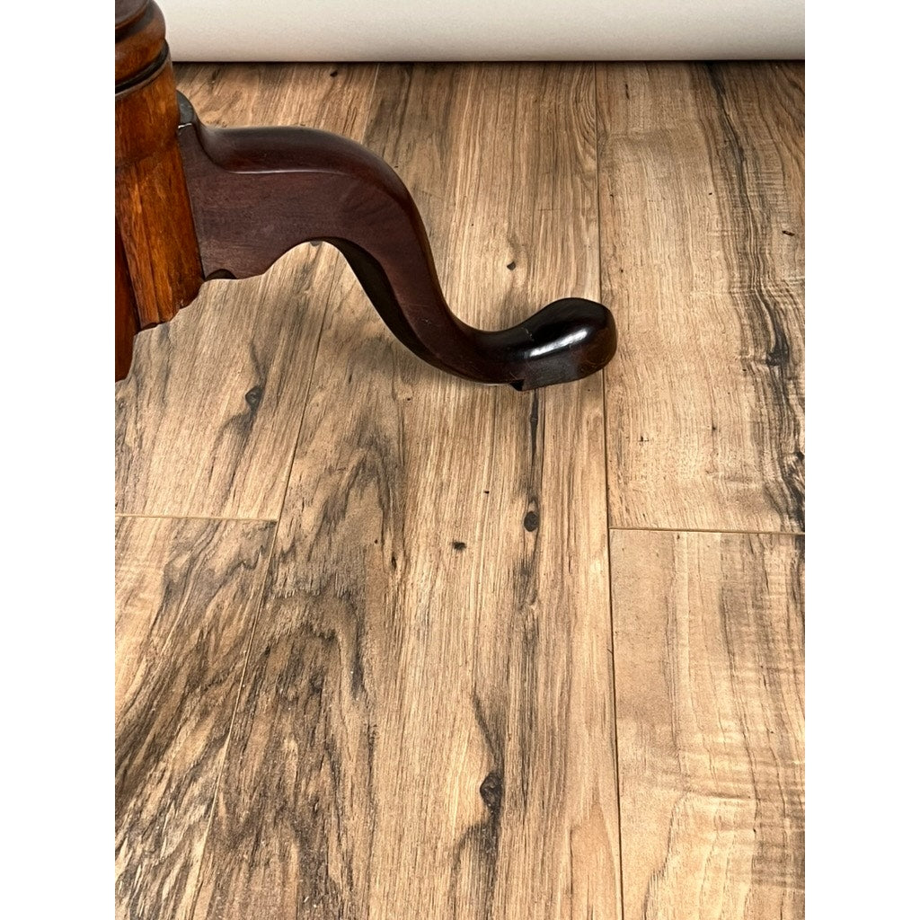 A curved, dark mahogany leg from an Antique Queen Anne Mahogany Tilt Top Breakfast Table (late 18th century) rests on a light brown, wood-patterned floor with varied grain and color tones.