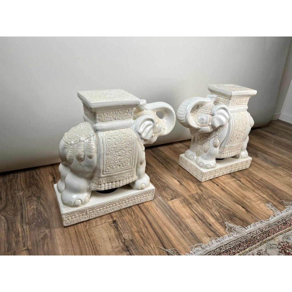 The Vintage Pair White on White Glazed Hollywood Regency Style Elephant Garden Seats feature ornate patterns and square flat tops, perfect as tables or stylish stands on a wooden floor by a white wall and patterned rug. Brand: Unknown.